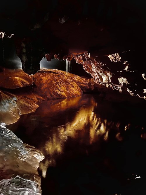 A dark cave with shimmering water reflecting warm tones and light filtering through rocky formations above.