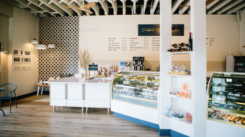Bright café interior featuring a display case of pastries, a coffee station, and modern decor with patterned walls.
