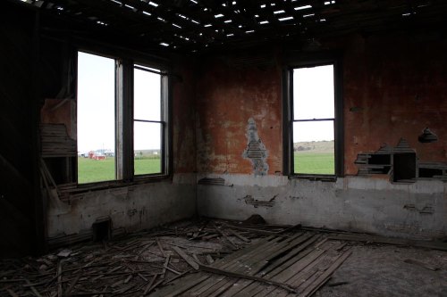 Interior of a dilapidated room with peeling walls, broken windows, and scattered wooden debris, overlooking a green field.