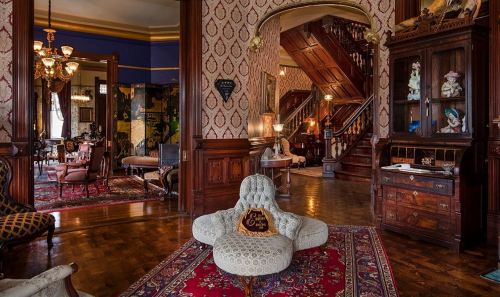 A vintage living room with ornate decor, a plush chair, and a grand staircase in the background. Warm, inviting atmosphere.