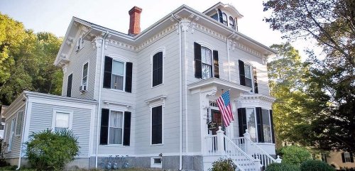 A large, historic white house with black shutters and an American flag, surrounded by greenery.