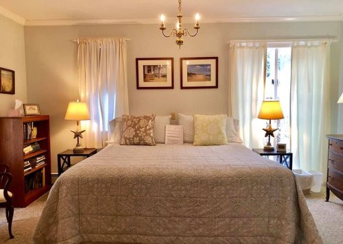 Cozy bedroom with a large bed, decorative pillows, two lamps, and soft lighting from windows and a chandelier.