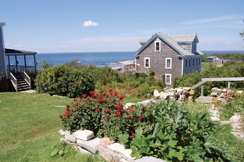 A coastal scene featuring a house by the sea, surrounded by greenery and vibrant red flowers under a clear blue sky.