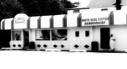 Black and white image of a diner named "White Rose System Hamburgers" with a striped awning and a person standing outside.