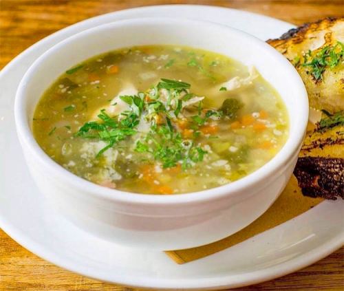 A bowl of soup with vegetables and herbs, served with a slice of toasted bread on the side.