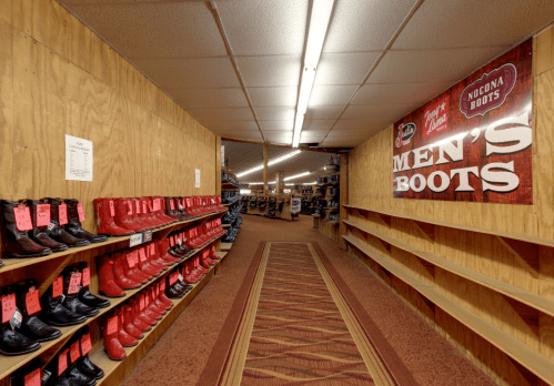 A spacious store aisle lined with shelves of men's boots, featuring red boots and a large "Men's Boots" banner.