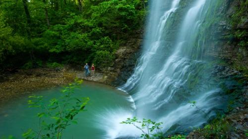 Two people stand near a waterfall surrounded by lush greenery and a serene pool of water below.