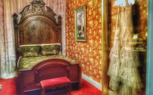 A vintage bedroom featuring a wooden bed, ornate wallpaper, and a lace dress hanging nearby.
