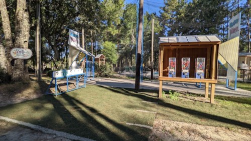 A playground scene featuring a slide, gumball machines, and trees in a sunny outdoor setting.