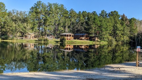 A serene lake reflecting trees and cabins on the shore, surrounded by lush greenery under a clear blue sky.