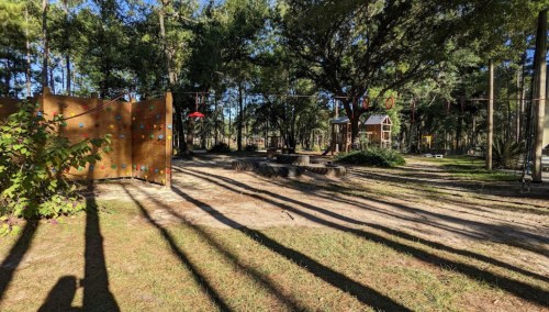 A playground in a wooded area featuring climbing structures, a rock wall, and long shadows from trees.