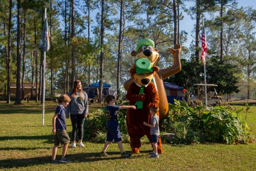 A costumed bear character poses with children in a grassy area, surrounded by trees and flags.