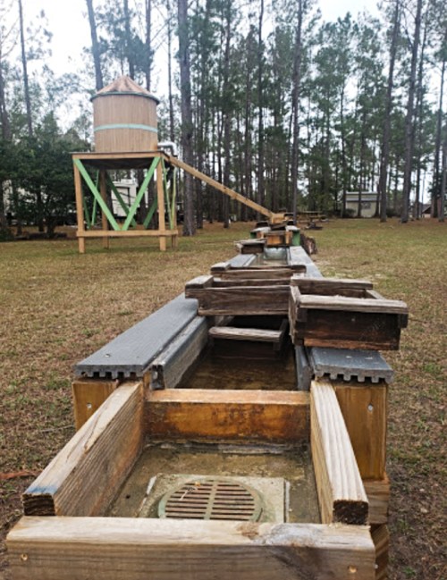 A wooden water trough with a water tower in the background, surrounded by trees in a grassy area.