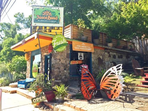 A colorful juice shop with a butterfly sculpture outside, surrounded by greenery and a welcoming "Open" sign.