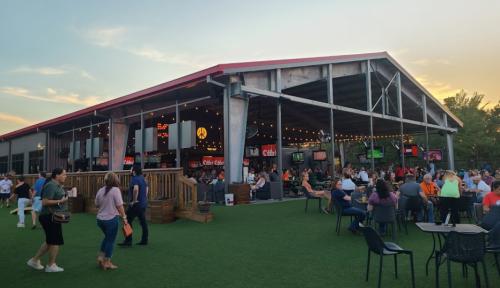 A lively outdoor venue with people socializing, dining, and enjoying the evening under string lights and a sunset sky.