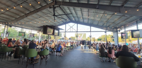 A lively outdoor venue with people seated at tables, string lights overhead, and screens displaying events.