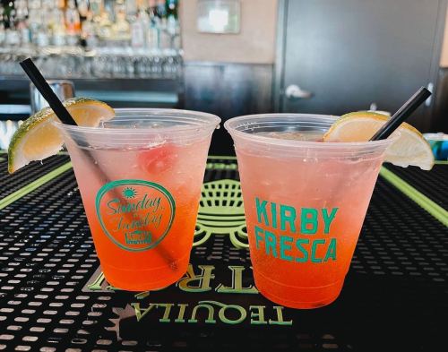 Two colorful cocktails in clear cups with lime and lemon slices, sitting on a bar counter.