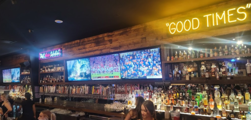 A lively bar scene with multiple TVs showing sports, shelves lined with various liquor bottles, and a neon sign reading "Good Times."