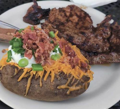 A loaded baked potato topped with cheese, bacon, and green onions, served with grilled meat on a plate.