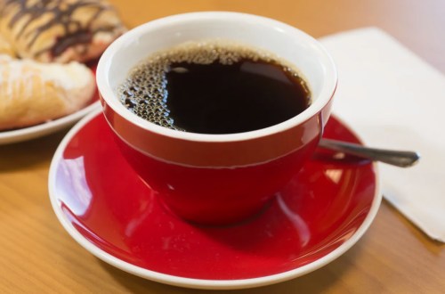 A red cup filled with black coffee on a matching saucer, next to a plate of pastries.