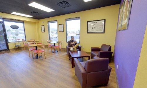Cozy café interior with yellow and purple walls, featuring tables, chairs, and a person sitting in a lounge chair.