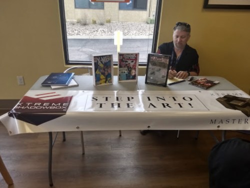A person sits at a table with comic books and a banner that reads "Step Into The Art!" in a casual setting.