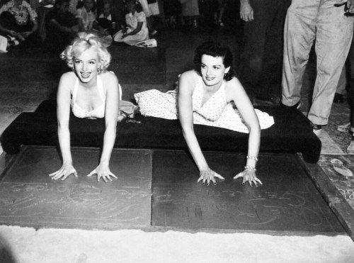 Marilyn Monroe and another actress press their hands into cement at a Hollywood event, smiling and posing for the camera.