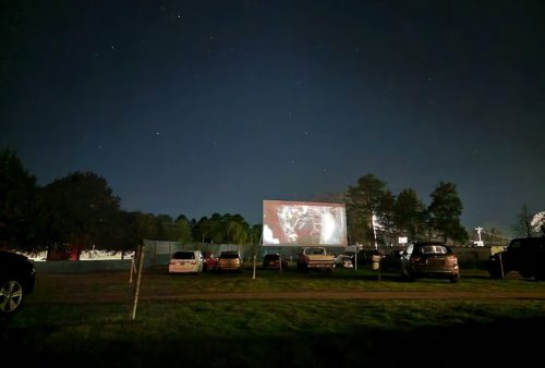 A drive-in movie theater at night, with cars parked in front of a large screen displaying a film under a starry sky.