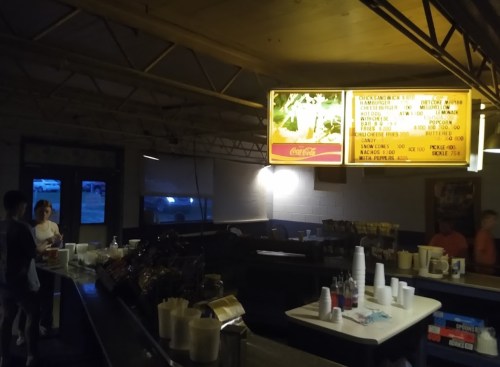 A dimly lit concession stand with a menu board, tables, and people in the background.