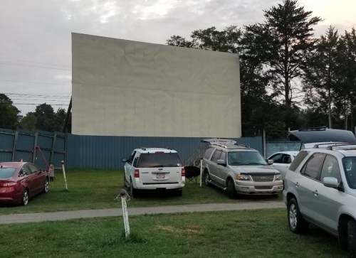 A blank outdoor movie screen stands behind parked cars in a grassy area, surrounded by trees and a cloudy sky.