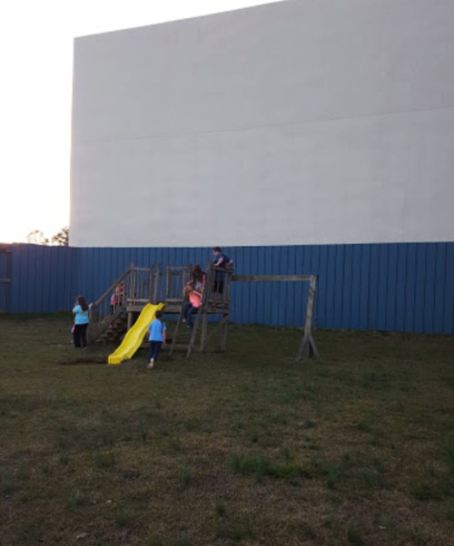 Children playing on a wooden playground with a slide, set against a large white wall at sunset.