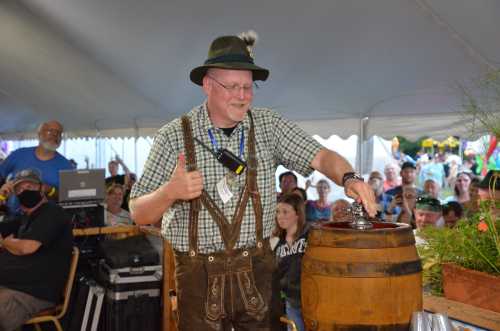 A man in traditional attire gives a thumbs up while pouring from a wooden barrel at a festive outdoor event.