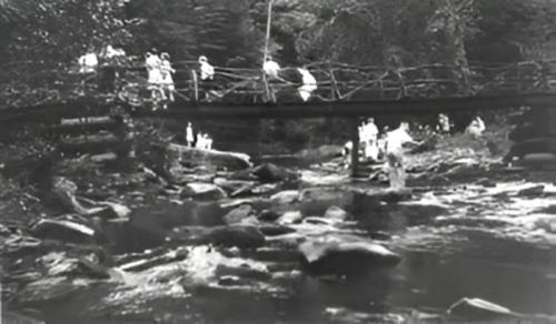 A black and white photo of a wooden bridge over a rocky stream, with people walking across and surrounding the area.
