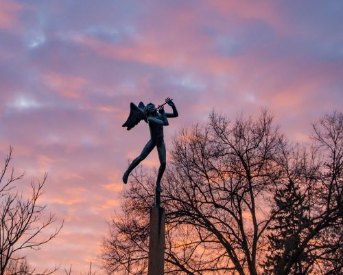 A bronze statue of an angel playing a flute against a colorful sunset sky, with silhouetted trees in the foreground.