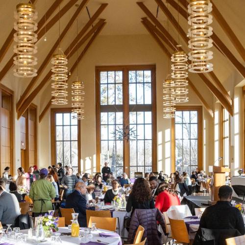 A spacious dining hall filled with people enjoying a meal, featuring large windows and modern lighting fixtures.