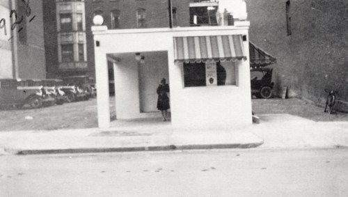 A black-and-white photo of a small building with a striped awning, a woman standing outside, and vintage cars in the background.