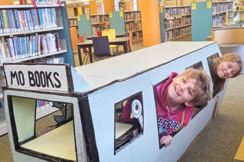 Two children play inside a cardboard bookmobile in a library, smiling and enjoying their imaginative experience.