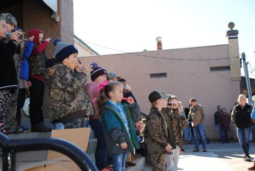 A group of children in camouflage and winter clothing stand on steps, some holding instruments, during an outdoor event.