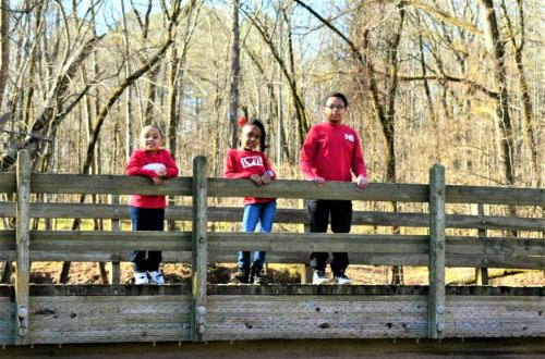 Three children in red shirts stand on a wooden bridge surrounded by trees in a natural setting.