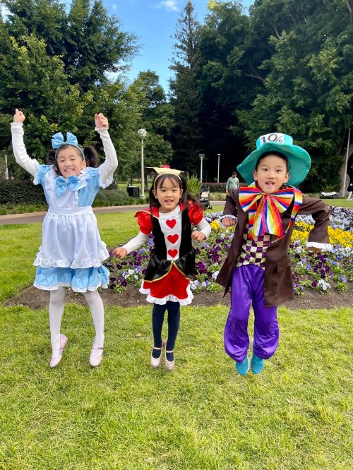 Three children in colorful costumes jump joyfully in a park, surrounded by flowers and greenery.