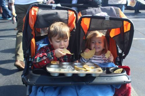 Two young children in a stroller enjoying snacks, with one eating a taco and the other smiling happily.