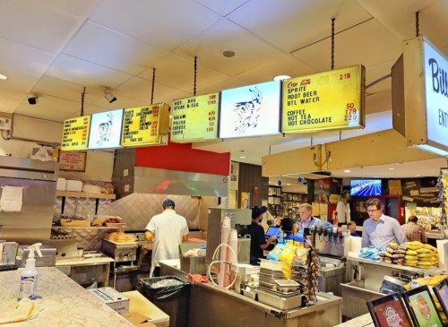 A busy diner with a menu board displaying food and drink prices, staff preparing orders in a vibrant kitchen setting.