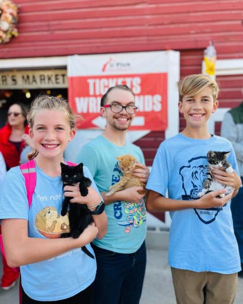 Three smiling kids hold kittens outside a market, with a festive backdrop and a sign for tickets in the background.