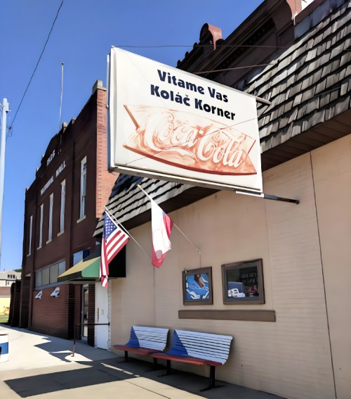 Sign for "Vitame Vas Kolač Korner" with a Coca-Cola logo, in front of a brick building and benches outside.