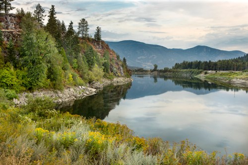 A serene river scene with lush greenery, rocky cliffs, and mountains reflected in calm water under a cloudy sky.