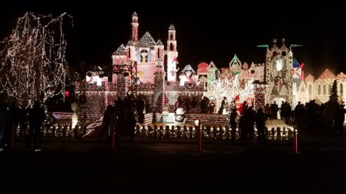 A festive display of colorful holiday lights illuminating a decorated house and surrounding area, with a crowd enjoying the scene.