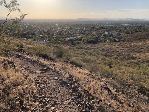 A rocky trail leads down a hillside, overlooking a sprawling valley and distant mountains under a clear sky.