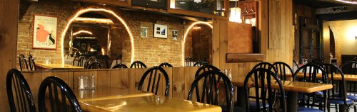 Cozy restaurant interior with wooden tables, black chairs, and brick walls, featuring mirrors and warm lighting.