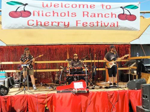 A band performs on stage at the Nichols Ranch Cherry Festival, with a colorful banner and festive decorations.