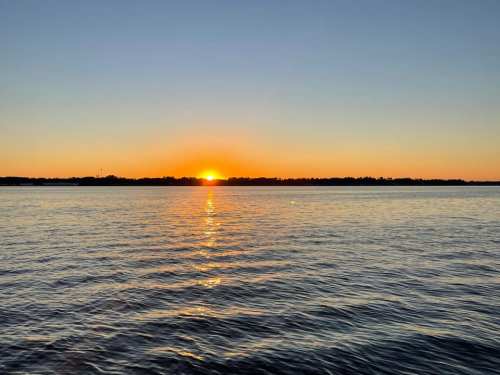 A serene sunset over calm water, with the sun dipping below the horizon and a clear sky.